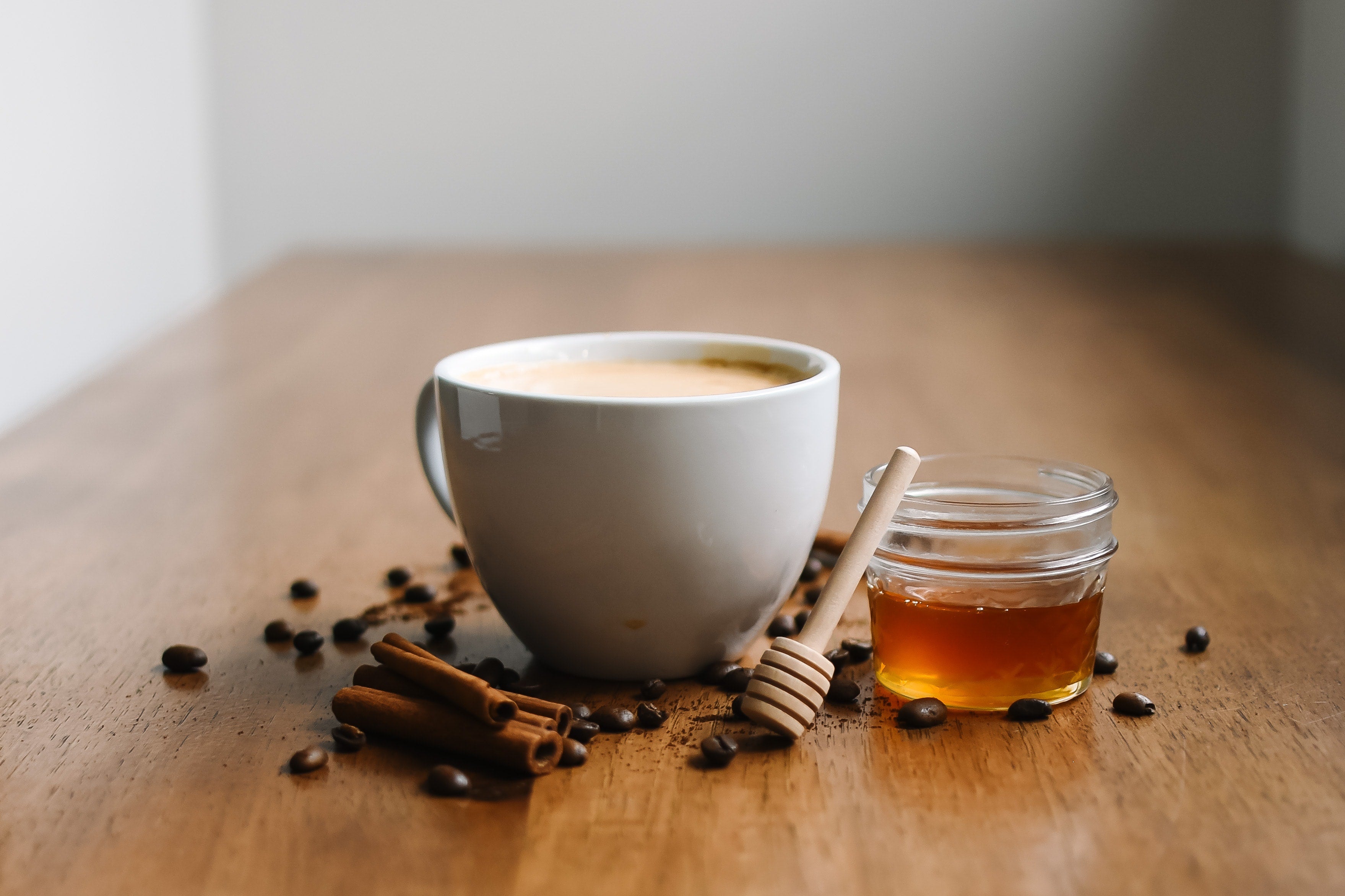 A warm cup of coffee with a jar of honey, honey dipper, cinnamon stick and scattered coffee beans on a wooden surface